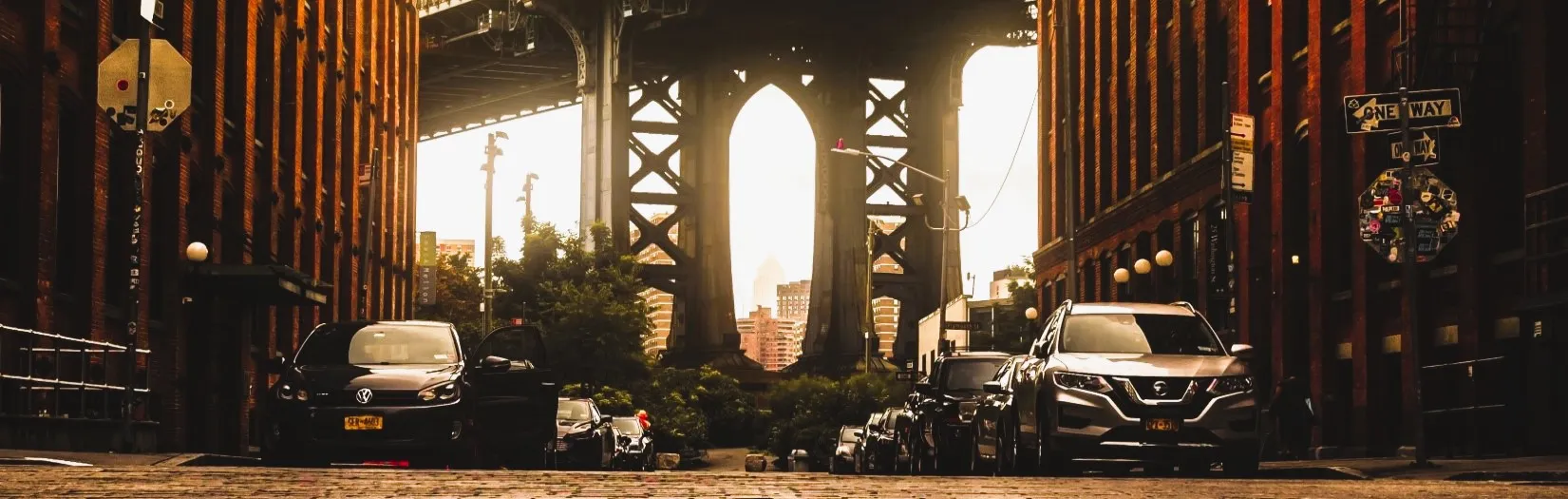 A panoramic view of the bottom of a huge bridge, could be Brooklyn, NY. There are many cars parked on the side of the road. Our category of Car Stuff found at Ads4Dads.com