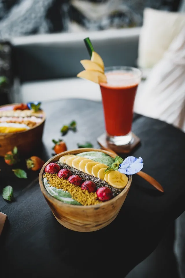 A bowl filled with pureed fruit and the top is decorated with fruits like bananas, cherries. There is also a mint leaf on the side with a small flower. In the background there is a healthy drink that appears to be pureed fruits and vegetables. Learn how to live a Healthy lifestyle here at Ads4dads.com