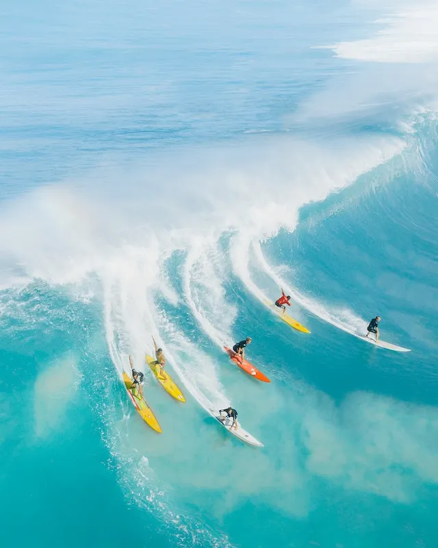 An arial view of six surfers all on the same wave. It's a beautiful blue with different colored surfboards. Surfing is more enjoyable with Jesus by your side. -Ads4dads.com