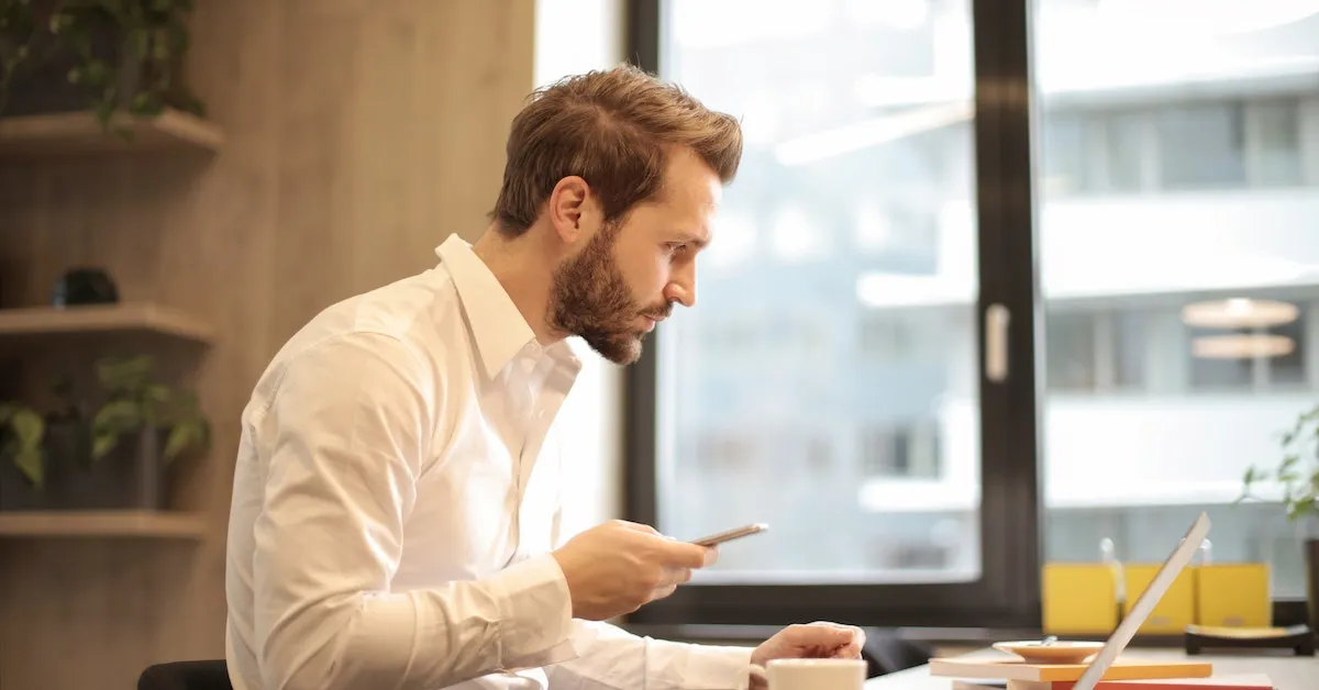 A man looking at a computer while holding his cell phone. There is a coffee cup and a breakfast plate in the background. Learn how to improve your morning routine in this article.