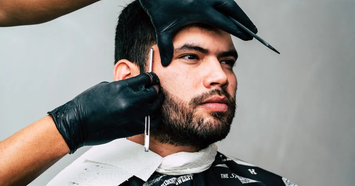 A close-up image of a man at a barbershop. He is having his beard shaped by a straight razor. Are you ready to save money and cut your own hair? Learn more here.