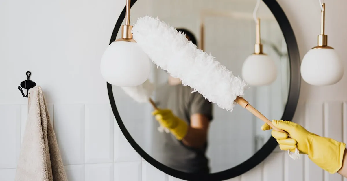 A person using a white dust wand to clean a mirror and lamp. Learn how to remove excessive dust in your home in this article. -Ads4Dads