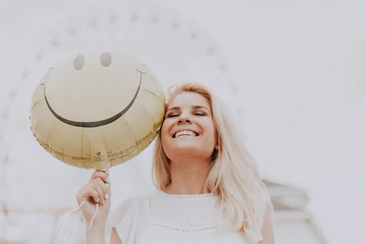 A pretty woman with blonde hair smiling in front of a ferris wheel. She is also holding a large yellow balloon that has a smiley face on it. She has lots of courage and is strong in hope, just like King David has. learn more in this latest article.