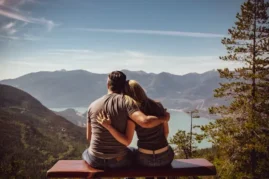 A married couple embracing while looking out into the distance of large mountains and a beautiful lake.
