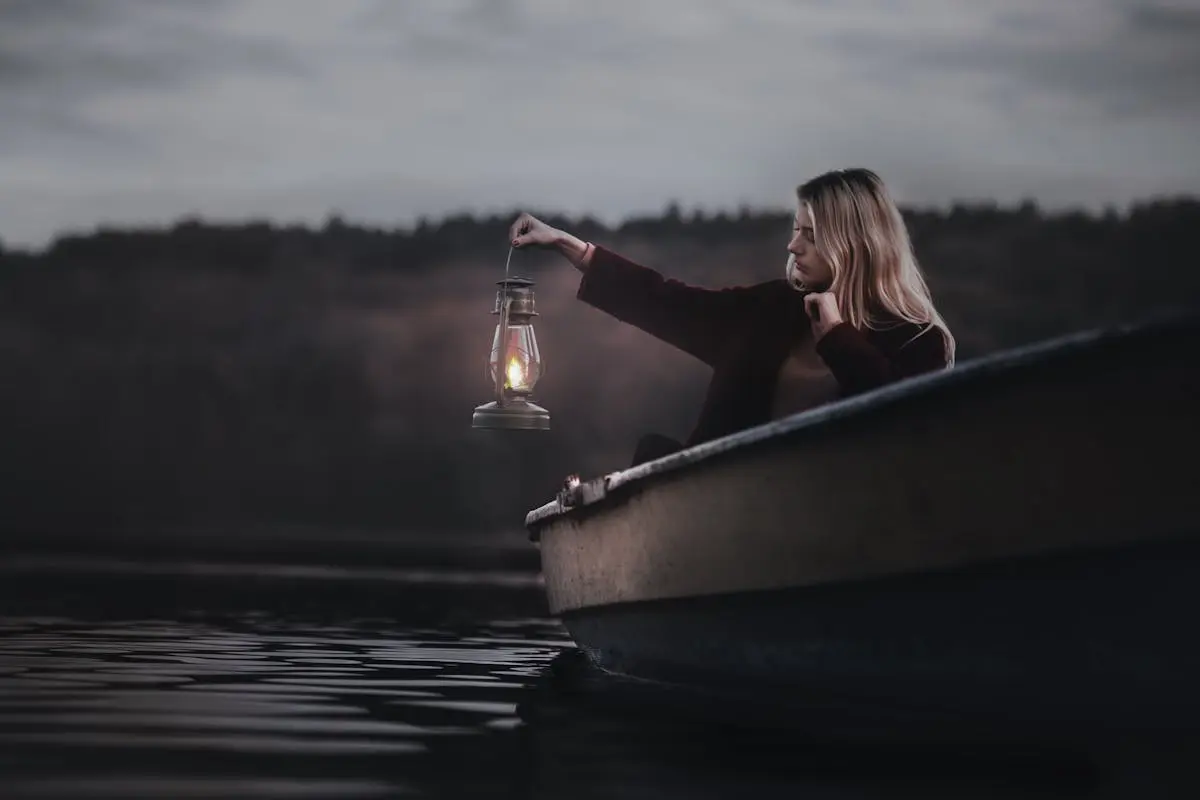 A woman holding a lantern while sitting in a boat. She is in the middle of a lake just before dark.