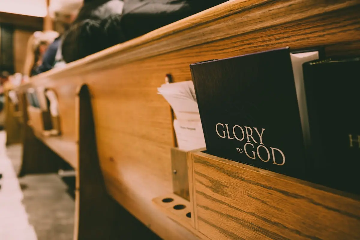 The back side view of a church pew with a Bible in the wooden book holder. The words on the front cover of the Bible read: Glory to God