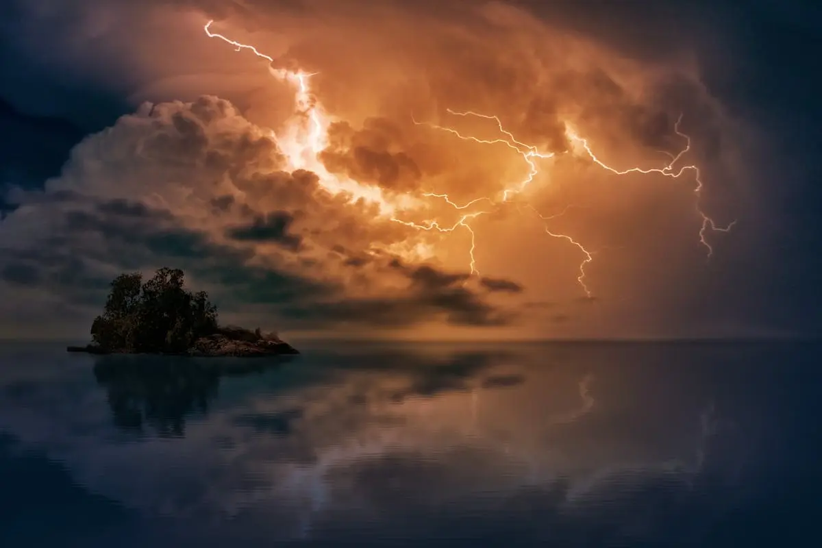 A small island on the ocean with a fierce storm rolling in. There is a lightning filling up the sky like fire behind the clouds.